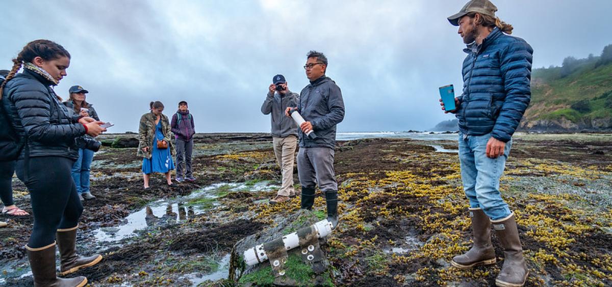 Francis Chan stands in tide pool on the Oregon Coast, with ocean sensor in foreground and a group of people gathered around. 