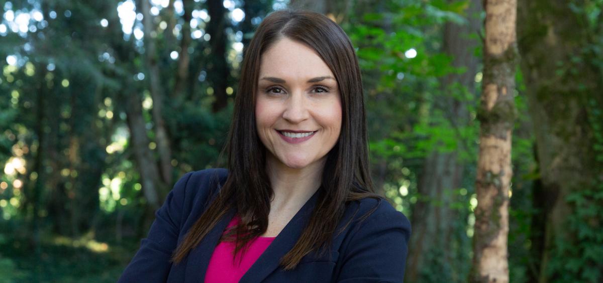 Headshot of Oregon Secretary of State Shemia Fagan in navy blazer and pink blouse and standing in front of a forest background