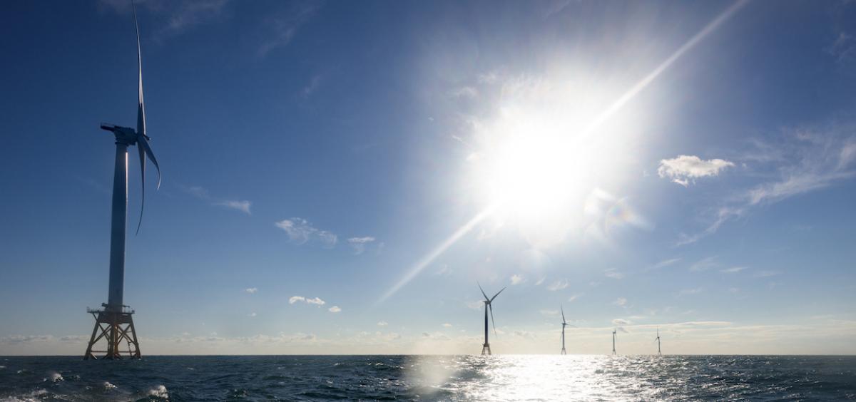 Image shows a series of 5 massive offshore wind turbines in open ocean under a glaring sun; the first turbine is close by in the foreground and the others look progressively smaller as they are farther away.