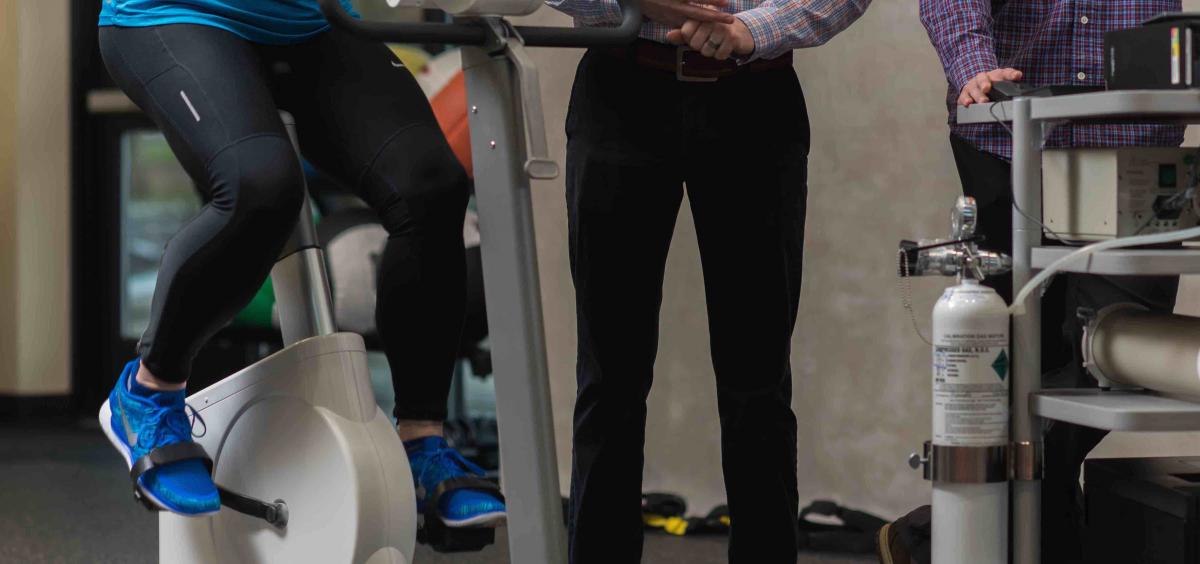 Image of a woman waist-down riding an exercise bike while two researchers stand nearby