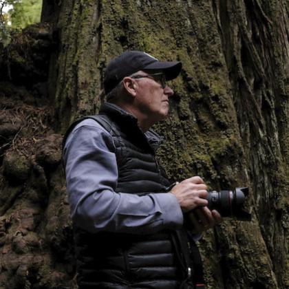 Photographer Chris Johns appears in a black vest and baseball cap, facing away from the camera to the right amid a stand of big trees