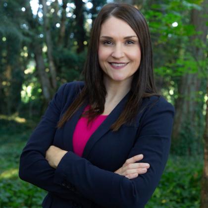 Headshot of Oregon Secretary of State Shemia Fagan in navy blazer and pink blouse and standing in front of a forest background