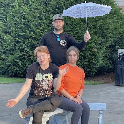 Image from a Twelfth Night rehearsal shows two people seated on a bench, one with arms outstretched in a comical gesture, while a third stands behind holding a white parasol over their heads.