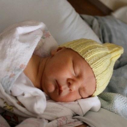 Stock image of infant, wrapped in white blanket and with a yellow knit beanie on its head