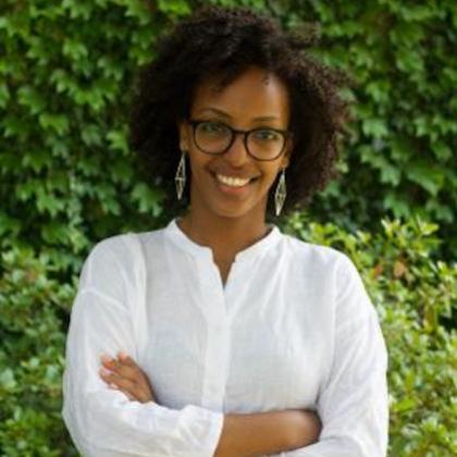 Image of Adom Getachew, wearing glasses, dangly earrings and a white button-up blouse against a background of green leaves
