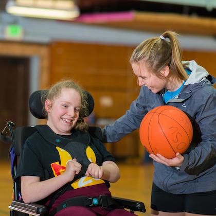 Image shows a volunteer holding a basketball and smiling with a student participant in a wheelchair. Both are in a gymnasium, participating in OSU's IMPACT program.