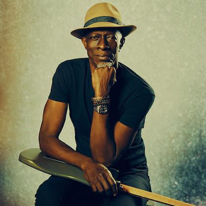 Musician Keb' Mo' looks directly at the camera, sitting up with his guitar atop his lap and resting his chin on his fist. He wears a tan fedora-style hate and a dark t-shirt and pants. He looks very cool.