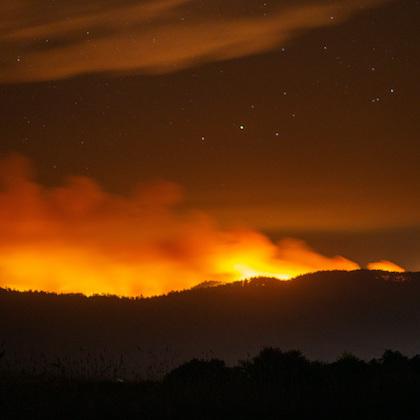 Image of red-orange glow of wildfire and smoke creeping over a darkened hillside at night