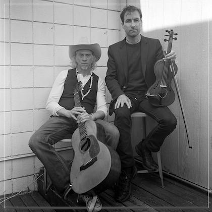 Black & white image of musicians Jimbo Mathus and Andrew Bird; Mathus leans back in his chair, holding a guitar between his legs, while Bird sits on a taller stool, resting a violin upright on his thigh. 
