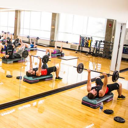 An instructor leads students in a Body Pump class at OSU; students are arrayed about a fitness room in front of a wall of mirrors, lying back on stacks of risers and raising barbells over their heads.