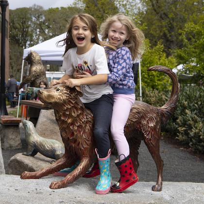 Two children laughing atop the lifesize bronze sculpture of a dog outside OSU's veterinary college at Pet Day 2022.