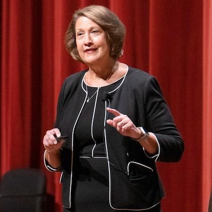 Ruth Watkins speaks onstage in front of the orange curtain at OSU's University Day, wearing a black dress with white piping and a matching blazer.