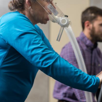 Image of a woman with an plastic mask on her face & tube extending from it while she rides an exercise bike