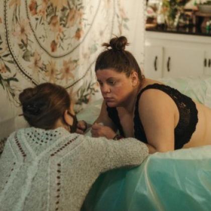 A dark-haired woman in labor leans against the edge of a water-birth tub while a birthing coach crouches next to her. 