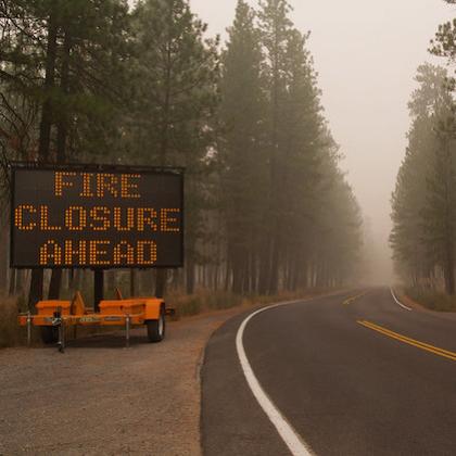 In a smoky forest, a large road sign on the left of a two-lane road says "Fire Closures Ahead." Image was taken during the 2020 wildfires in Oregon. 