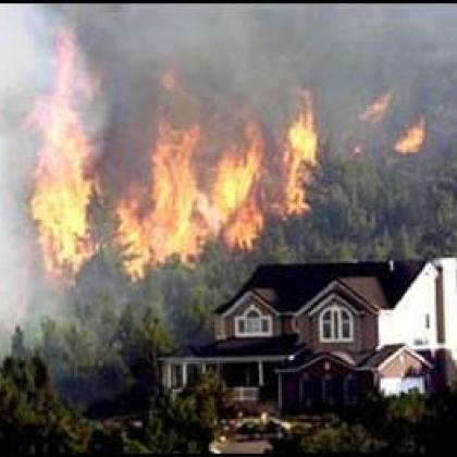Masive flames billow up among the pine trees behind a large house on the edge of the forest.